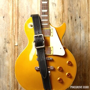 May include: A gold electric guitar with a black leather strap. The guitar has a white pickguard and gold hardware. The strap has a buckle and stitching.