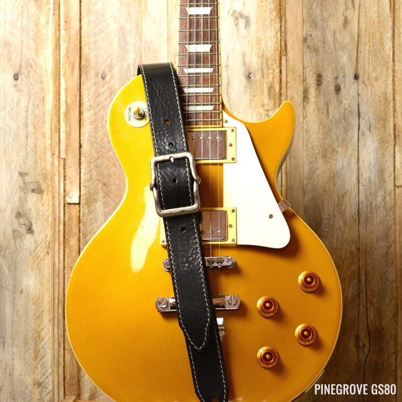 May include: A gold electric guitar with a black leather strap. The guitar has a white pickguard and gold hardware. The strap has a buckle and stitching.