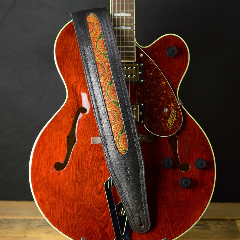 May include: A black leather guitar strap with a colorful paisley pattern. The strap is attached to a red hollow-body electric guitar with a Gretsch logo.