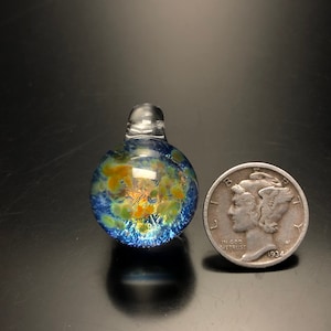 May include: A clear glass pendant with a blue and orange floral design. The pendant is round with a small loop at the top. A silver coin is next to the pendant for size comparison.