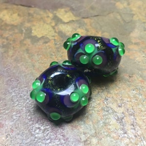 May include: Two dark blue glass beads with a speckled gold finish. The beads are decorated with green and purple dots.