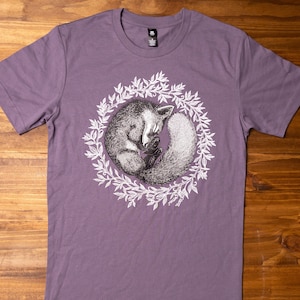 May include: A purple t-shirt with a graphic of a sleeping fox curled up inside a wreath of leaves. The fox is black and white and the wreath is white.