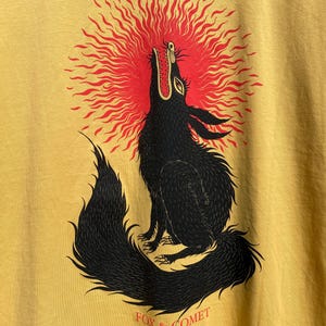 May include: Mustard yellow t-shirt featuring a black illustration of a howling wolf with a red sunburst behind its head. The text "FOX & COMET" is printed in red below the wolf.