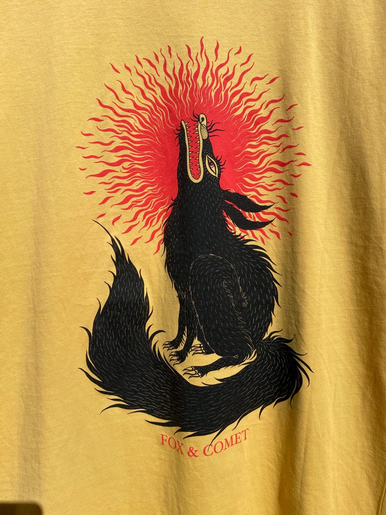 May include: Mustard yellow t-shirt featuring a black illustration of a howling wolf with a red sunburst behind its head. The text "FOX & COMET" is printed in red below the wolf.