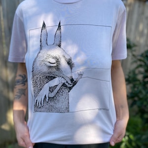May include: White t-shirt with a black and white illustration of a creature with large ears holding a human arm. The text "MIKE BURKE" is printed in the bottom right corner of the illustration.