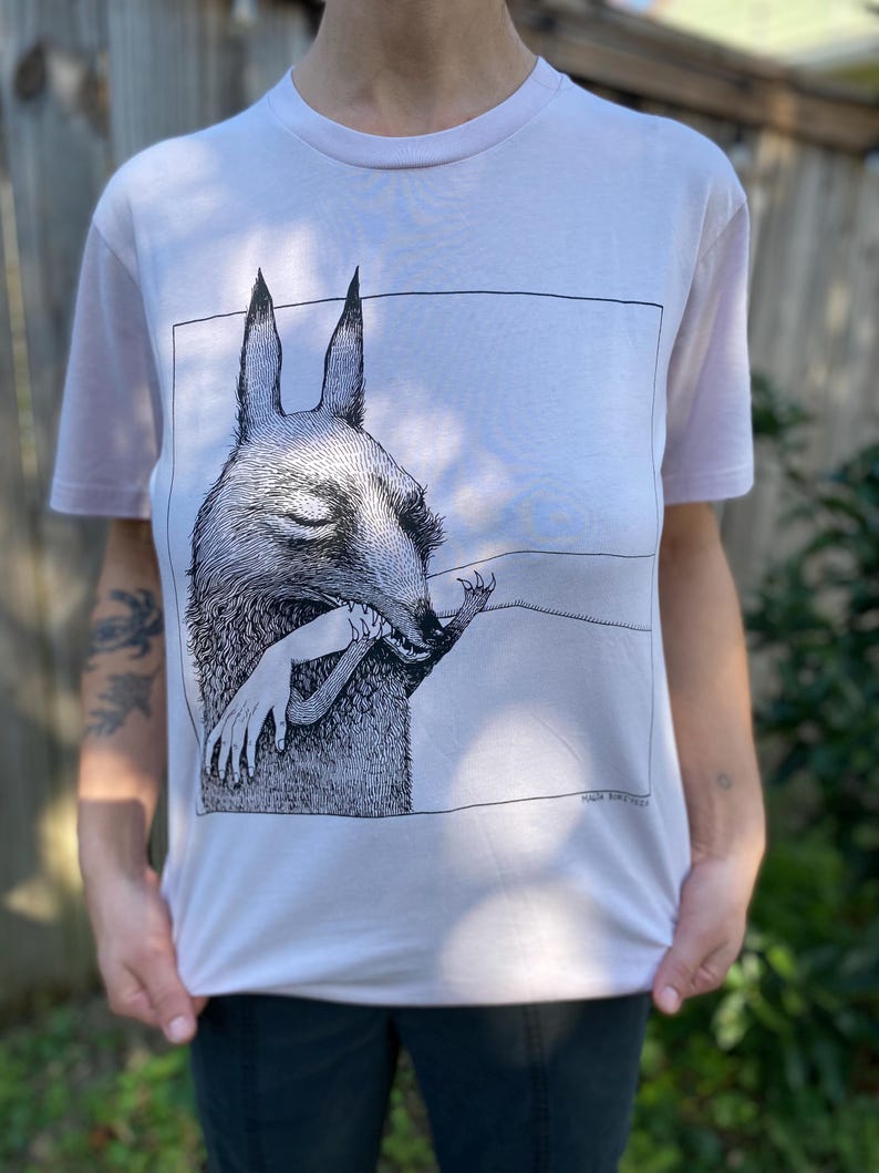 May include: White t-shirt with a black and white illustration of a creature with large ears holding a human arm. The text "MIKE BURKE" is printed in the bottom right corner of the illustration.