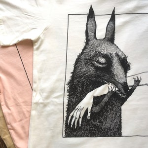 May include: A white t-shirt with a black and white illustration of a wolf biting a hand. The illustration is framed in a black square with the word "MAGMA" in the bottom right corner.