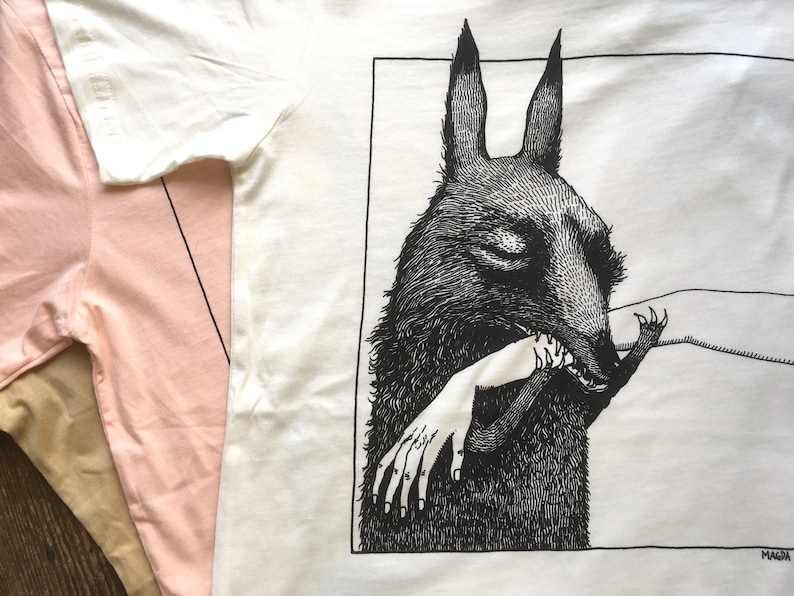 May include: A white t-shirt with a black and white illustration of a wolf biting a hand. The illustration is framed in a black square with the word "MAGMA" in the bottom right corner.
