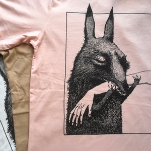 May include: A pink t-shirt with a black and white illustration of a creature with large ears biting a hand. The illustration is framed in a black rectangle. The text "MAGDA BORBA" is at the bottom right corner of the image.