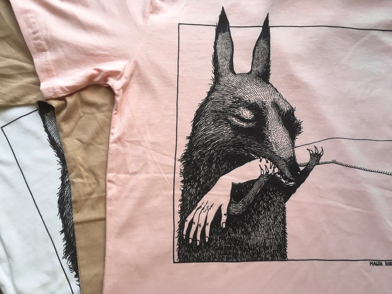 May include: A pink t-shirt with a black and white illustration of a creature with large ears biting a hand. The illustration is framed in a black rectangle. The text "MAGDA BORBA" is at the bottom right corner of the image.