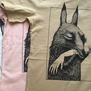 May include: Three t-shirts with a black and white illustration of a wolf-like creature biting a hand. The t-shirts are in beige, pink, and white.
