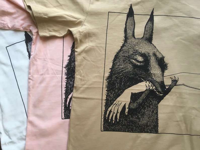 May include: Three t-shirts with a black and white illustration of a wolf-like creature biting a hand. The t-shirts are in beige, pink, and white.
