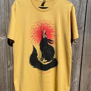 May include: Mustard yellow t-shirt featuring a black wolf howling at a red sunburst design. The text "FOX & COMET" is printed below the wolf. The shirt is hanging on a black hanger.