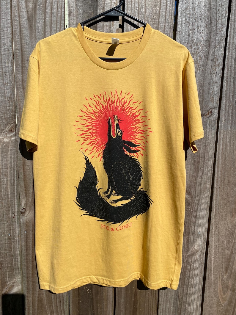 May include: Mustard yellow t-shirt featuring a black wolf howling at a red sunburst design. The text "FOX & COMET" is printed below the wolf. The shirt is hanging on a black hanger.