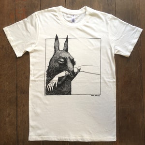 May include: White t-shirt with a black and white illustration of a creature with large ears biting a hand. The text "FRANK STELLA" is printed below the illustration.