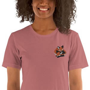 May include: A short-sleeved, crew-neck t-shirt in a dusty rose colour. It features an embroidered design of two foxes embracing, in orange, black, and white. The t-shirt is made of a soft material.