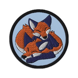 Könnte beinhalten: Gestickter Aufnäher mit zwei roten Füchsen, die sich aneinanderkuscheln, einer liegt auf dem anderen, mit blauen Akzenten. Die Füchse befinden sich auf einem hellblauen Hintergrund mit einem schwarzen runden Rand.