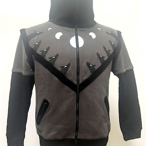Op de afbeelding: Een grijs ritsjack met zwarte mouwen en een zwarte mock neck. De voorkant van het jack heeft een zwart-wit ontwerp van wolven die naar de maan huilen.