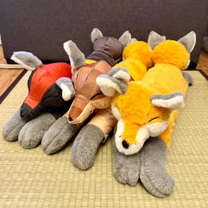 May include: Three plush fox toys lying side by side. The left fox is red and black, the middle fox is brown and tan, and the right fox is bright orange and white. Each fox has gray paws, pointed ears, and a soft, textured fabric finish.