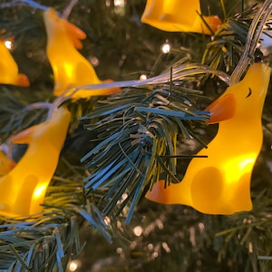 May include: String of yellow dog-shaped lights, illuminated and strung on a clear wire. The lights are set against a backdrop of green Christmas tree branches, creating a festive and whimsical display.