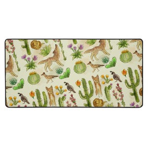 May include: Rectangular desk mat with a desert-themed pattern. The design features illustrations of cacti, coyotes, roadrunners, and desert flowers in green, brown, orange, and purple on a cream background. The mat has a black border.