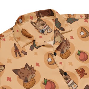 May include: A button-down shirt with a repeating pattern of cartoon wolf characters, pumpkins, apples, and fall-themed elements. The shirt is a light orange color with a collar and black buttons.