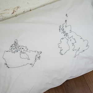 Puede incluir: Una funda de almohada blanca con contornos bordados en negro de los mapas de Canad&aacute; y el Reino Unido.