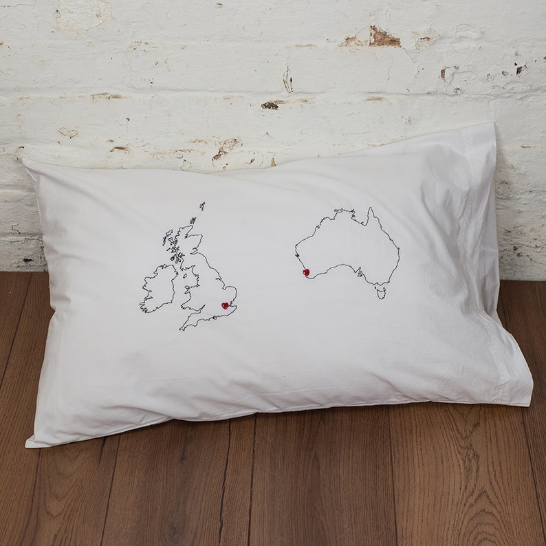 Puede incluir: Una funda de almohada blanca con contornos bordados del Reino Unido y Australia. Corazones rojos marcan ubicaciones en ambos mapas.