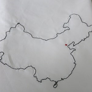 Puede incluir: Una tela blanca con un contorno bordado en negro del mapa de China. Un peque&ntilde;o punto rojo se encuentra en el centro del mapa.