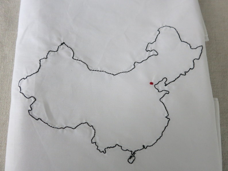 Puede incluir: Una tela blanca con un contorno bordado en negro del mapa de China. Un peque&ntilde;o punto rojo se encuentra en el centro del mapa.