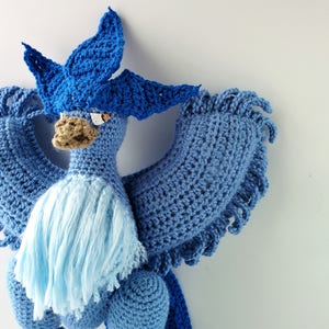 May include: A blue crocheted bird with a long, flowing tail and a blue and white crocheted crest. The bird has a brown beak and yellow eyes.