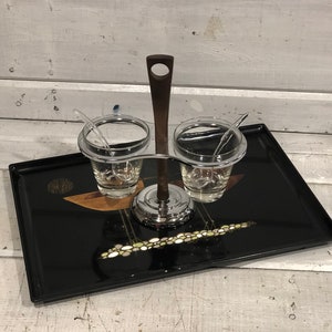 Könnte beinhalten: Ein schwarzes Lacktablett mit goldenen Akzenten trägt einen silbernen Gewürzhalter mit zwei Glasbehältern und Löffeln. Der Halter hat einen dunklen Holzgriff.