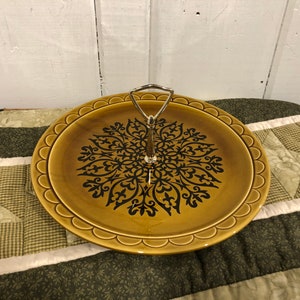 Puede incluir: Una bandeja de servir de cerámica marrón con un diseño floral negro y un asa de metal plateado. La bandeja tiene un borde festoneado.