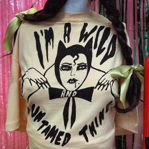 Könnte beinhalten: Cremefarbenes T-Shirt mit schwarzer Grafik einer vampirähnlichen Figur und dem Text "I'M A WILD AND UNTAMED THING". Das Shirt hat kurze Ärmel und wird auf einer Schaufensterpuppe präsentiert. Geflochtenes Haar mit grünen Schleifen ist sichtbar.