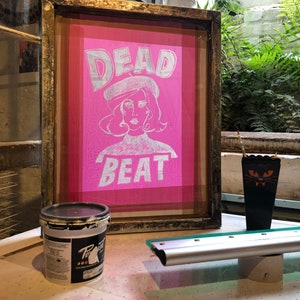 May include: A framed pink silkscreen print with the text "DEAD BEAT" in white lettering. A woman with a beret is depicted in the center of the print. A black paint can and a squeegee are also visible in the image.