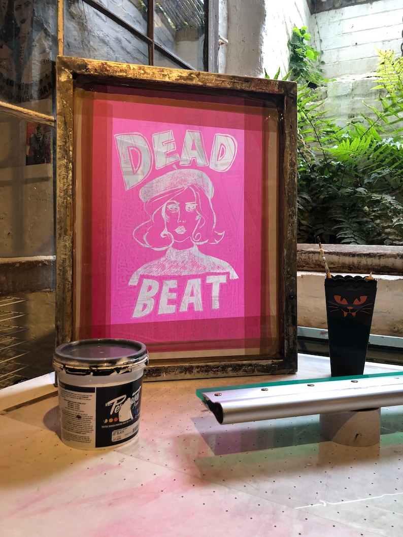 May include: A framed pink silkscreen print with the text "DEAD BEAT" in white lettering. A woman with a beret is depicted in the center of the print. A black paint can and a squeegee are also visible in the image.