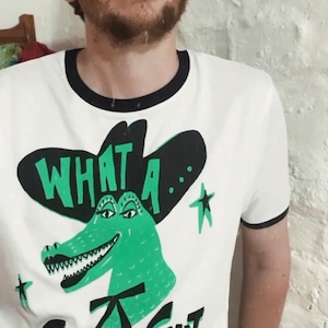 May include: White t-shirt with a black and green alligator wearing a black cowboy hat. The text "What a... Croc of Shit" is printed on the shirt.