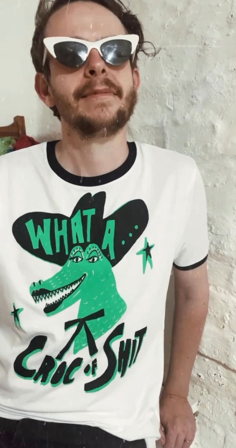May include: White t-shirt with a black and green alligator wearing a black cowboy hat. The text "What a... Croc of Shit" is printed on the shirt.