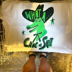 May include: White t-shirt with a green alligator wearing a black cowboy hat. The alligator has a toothy grin and the text "What a... Croc of Shit" is printed on the shirt.