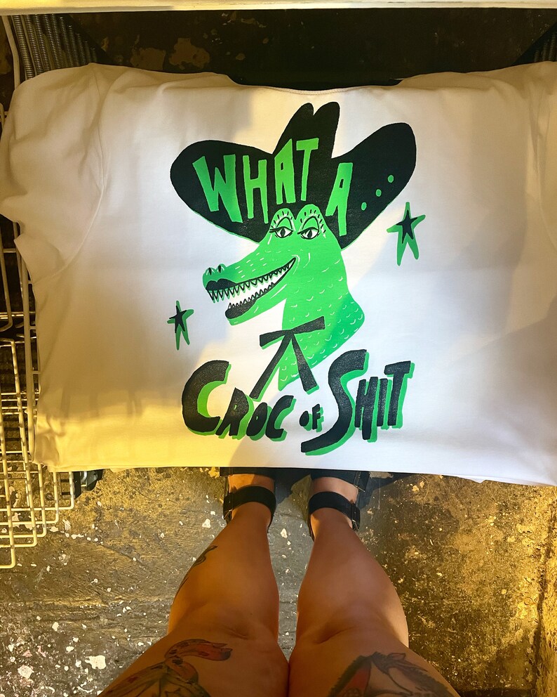 May include: White t-shirt with a green alligator wearing a black cowboy hat. The alligator has a toothy grin and the text "What a... Croc of Shit" is printed on the shirt.