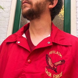 May include: Red nylon jacket with a collared neckline and snap closures. The jacket features an embroidered eagle design with the words "RECLAIM TASK FORCE" in gold lettering. A white undershirt is visible beneath the jacket.