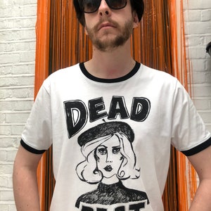 May include: A white t-shirt with a black ringer and a black and white graphic print of a woman wearing a beret with the text "DEAD BEAT" underneath.