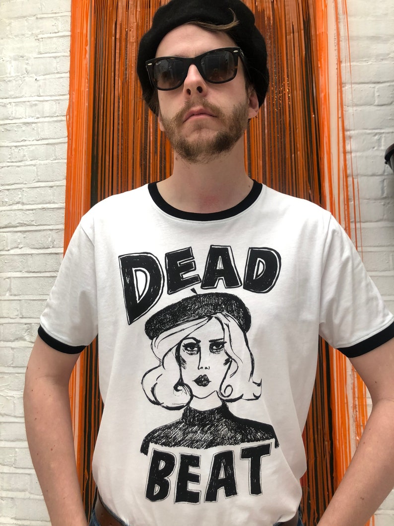 May include: A white t-shirt with a black ringer and a black and white graphic print of a woman wearing a beret with the text "DEAD BEAT" underneath.