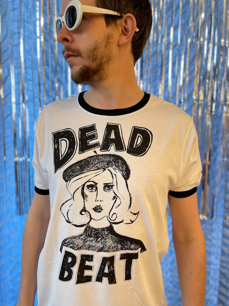 May include: White t-shirt with a black ringer and a black and white graphic print of a woman wearing a beret with the text "DEAD BEAT" underneath.