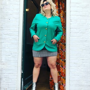 May include: A woman wearing a teal blazer with gold buttons, a black and white checked skirt, and silver ankle boots. She is standing in a doorway with a floral patterned curtain behind her.