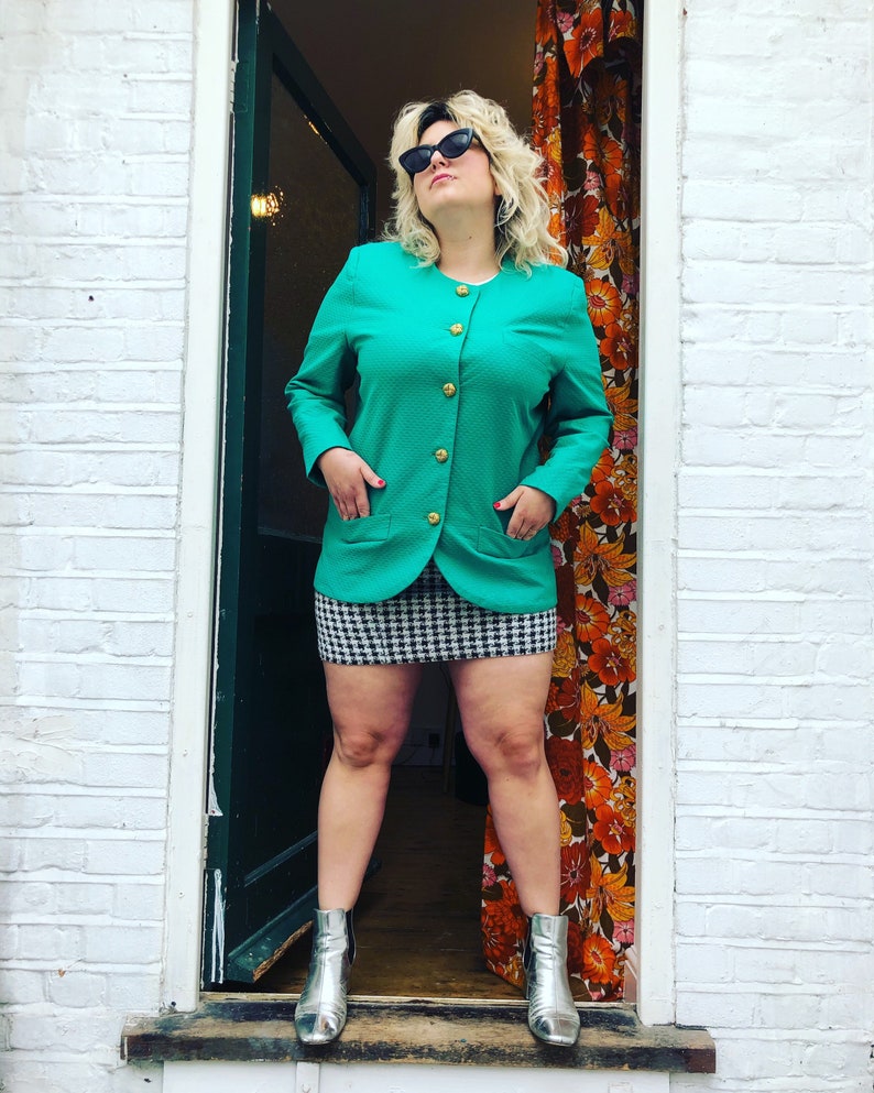 May include: A woman wearing a teal blazer with gold buttons, a black and white checked skirt, and silver ankle boots. She is standing in a doorway with a floral patterned curtain behind her.