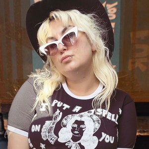 Könnte beinhalten: Ein schwarzes Ringer-T-Shirt mit weißem Saum an Ärmeln und Halsausschnitt, das einen weißen Grafikdruck einer Person mit Hut und den Text I PUT A SPELL ON YOU BECAUSE YOU'RE MINE zeigt, kombiniert mit einer weißen Cat-Eye-Sonnenbrille und einem schwarzen Hut.