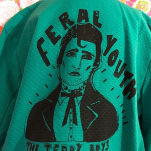 May include: A teal green jacket with a black graphic print of a man with the text "Feral Youth The Teddy Boys" on the back.
