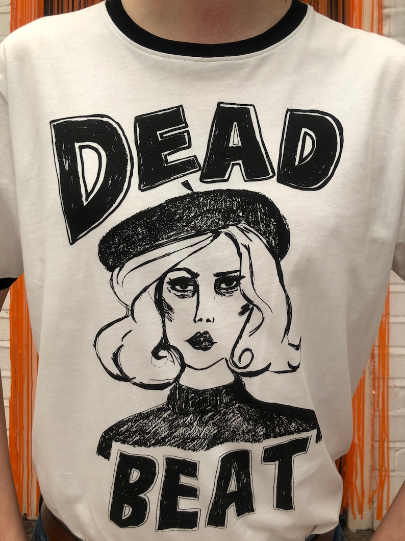 May include: White t-shirt with a black and white illustration of a woman wearing a beret and the text "DEAD BEAT" printed on it.