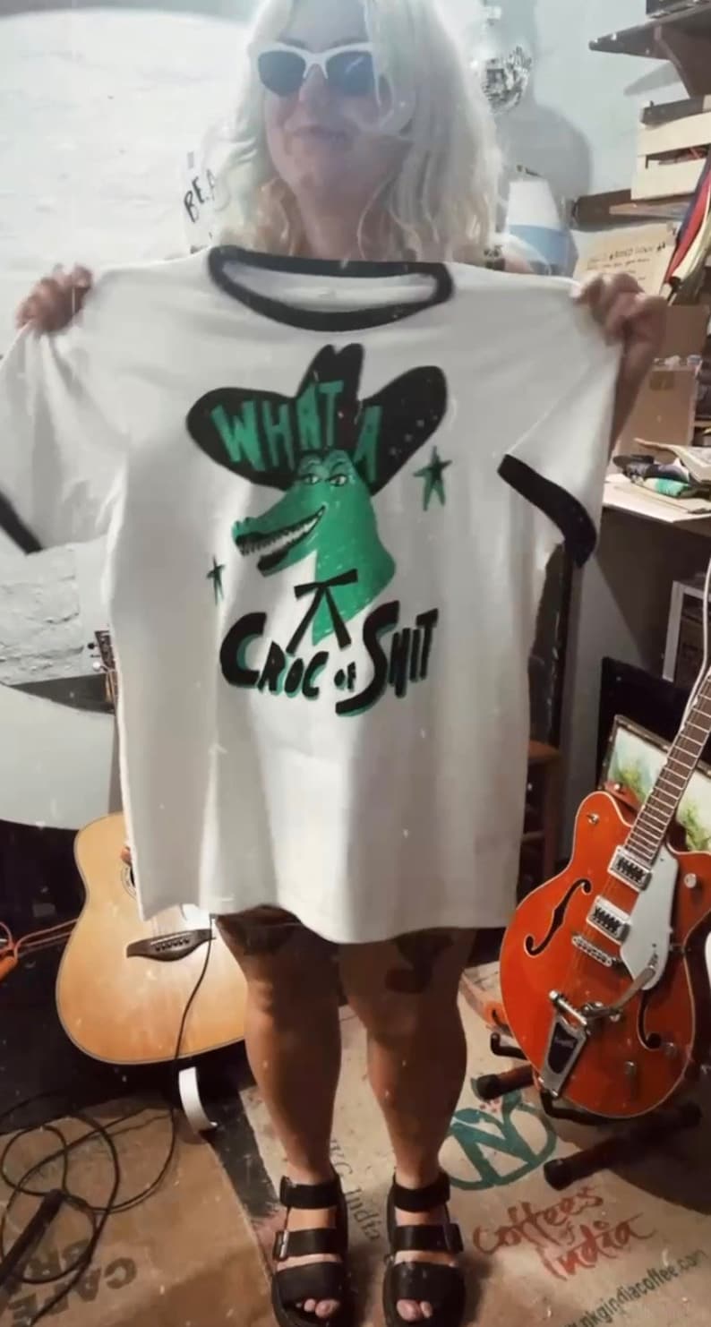 May include: White t-shirt with a green alligator wearing a cowboy hat and the text "What a Croc of Shit".